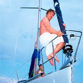 Man with towel around waist sitting on sailboat's railing, woman reading in background