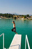 Boy jumping from diving platform into Wörthersee (biggest lake of Carinthia), Hotel Schloss Velden (served as the location for the TV series Ein Schloss am Wörthersee) in background, Strandbad, Velden, Carinthia, Austria