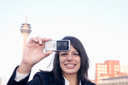 Young business woman making a selfportrait with mobile phone, in front of the city skyline, Media Harbour, Düsseldorf, harbor, television tower, Zollhof, architecture of Frank O.Gehry, state capital of NRW, North-Rhine-Westphalia, Germany