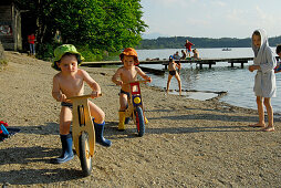 two boys with sunhat on juvenile bicycle, beach of lake Hartsee, Chiemgau, Upper Bavaria, Bavaria, Germany