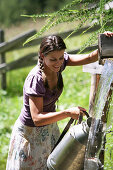 Woman filling milk can with water of a mountain stream, Heiligenblut, Hohe Tauern National Park, Carinthia, Austria