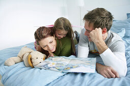 Junge Familie liegt auf einem Bett und liest ein Buch, München, Deutschland