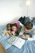Junge Familie liegt auf einem Bett und liest ein Buch, München, Deutschland