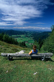 Young woman sitting on bench near alpine hut Kohleralm, view to Inzell, Kohler-Alm, Chiemgau range, Chiemgau, Bavarian foothills, Upper Bavaria, Bavaria, Germany