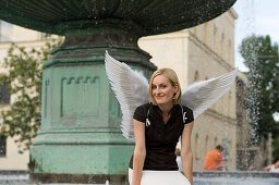 Frau mittleren Alters mit Engelsflügeln sitzt am Brunnen bei der Universität, München, Bayern, Deutschland