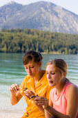Two young women sitting at lake Walchensee while eating, Bavaria, Germany