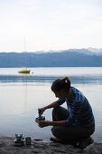 Eine junge Frau kocht Tee auf einem Gaskocher am Walchensee, Oberbayern, Bayern, Deutschland