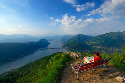 Frau mittleren Alters sitzt auf einer Bank, Luganer See im Hintergrund, Monte San Giorgio, Tessin, Schweiz