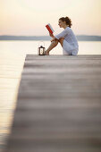 Woman sitting on jetty at lake Starnberg while reading a book, Ambach, Bavaria, Germany