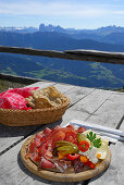 South Tyrolean snack with South Tyrolean speck, bacon, salami, peperoni, pepperoni, Kaminwurzen, cheese and Schuettelbrot, Dolomites in background, terrace of hut Radlseehuette, Sarntaler Alpen, Sarntal range, South Tyrol, Italy