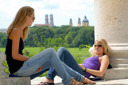 Zwei junge Frauen sitzen unter dem Monopteros mit Blick auf Frauenkirche und Theatinerkirche unscharf im Hintergrund, Englischer Garten, München, Oberbayern, Bayern, Deutschland
