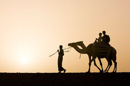 Ein Mann, Beduine führt zwei Kamele mit Touristen, Mutter mit zwei Kinder, Sonnenuntergang, Wüste von Marsa Alam, Rotes Meer Ägypten