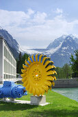 Rad einer Turbine bei Kraftwerkszentrale mit Staumauer des Albigna-Stausees und schneebedeckte Berge im Hintergrund, Peltonturbine, Wasserkraftwerk bei Löbbia, Bergell, Graubünden, Schweiz