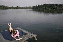 Young woman lying on a jetty while reading a book, lake Starnberg, Bavaria, Germany