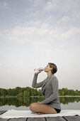 Junge Frau trinkt eine Flasche Wasser, sitzt auf einem Steg am Starnberger See, Bayern, Deutschland