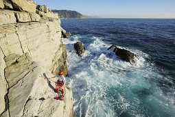 Woman standing on a cliff and rappelling, abseiling towards the spraying sea, cliff at the Mediterranean coast, Liguria, Italy