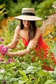 25 year old brunette woman in a garden settng wearing a dress and a straw hat cutting flowers for a bouquet