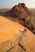 Frau klettert an roter Felswand, Sugarloaf im Hintergrund, Große Spitzkoppe, Namibia