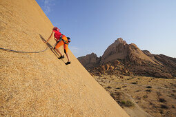 Frau klettert an roter Felswand, Große Spitzkoppe im Hintergrund, Große Spitzkoppe, Namibia