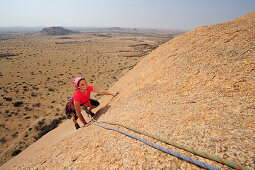 Frau klettert an roter Felsplatte über Savanne, Große Spitzkoppe, Namibia