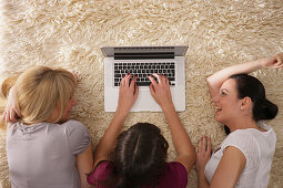Drei junge Frauen liegen mit einem Laptop auf einem Teppich, München, Bayern, Deutschland