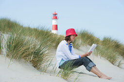 Woman wearing a red hut sitting at beach while reading a book, List Ost lighthouse in background, Ellenbogen, List, Sylt, Schleswig-Holstein, Germany