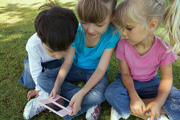 Three children sitting on grass, playing with video game, close-up