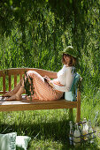 Frau sitzt mit einem Buch auf einer Gartenbank, Simssee, Bayern, Deutschland
