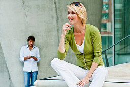 Woman sitting on step, man with mobile phone standing in background, HafenCity, Hamburg, Germany