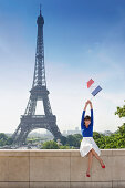 Woman holding a French flag sitting on a stone wall with the Eiffel Tower in the background, Paris, Ile-de-France, France