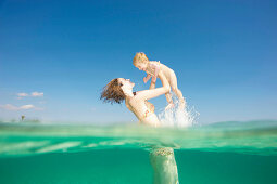 Mother lifting toddler up from water. Woman lifting young boy up in the air from crystal clear sea. Shot from both above and below water. Underwater shot.
