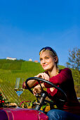 Junge Frau sitzt mit einem Glas Weißwein auf einem Traktor, Steiermark, Österreich