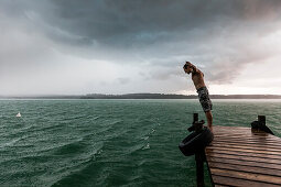 Young man on a jetty leaning into the wind, Lake Starnberg, Bavaria, Germany