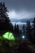Junge Frau mit Stirnlampe sitzt neben einem Zelt in einer Vollmondnacht, Bergell, Kanton Graubünden, Schweiz