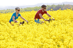 Two cyclists with electric bicycles between blooming canola fields, Tanna, Thuringia, Germany