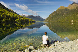 Spiegelung der Berge in Gunn, junge Frau sitzt am Ufer, Fiordland Nationalpark, Southland, Südinsel, Neuseeland