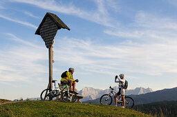 Mountain bikers resting beside a wayside cross, Winklmoosalm, Lofer Mountains in background, Chiemgau, Bavaria, Germany