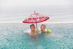 Vater und Sohn im Swimming Pool, roter Regenschirm, heftiger Regen, Monsun, Sturzregen, Meer, Überlaufpool, Luxus, tropischer Regen, Platzregen, Monsunregen, Monsun, Urlauber, Touristen, schwimmen, lachen, Junge 3 Jahre alt, Elternzeit in Asien, Europäer,