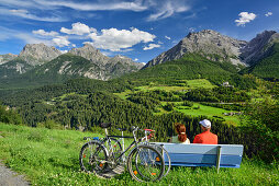 Two cyclists sitting on bench while enjoying view to castle of Tarasp and Sesvenna range, Scuol, Lower Engadin, Canton of Graubuenden, Switzerland