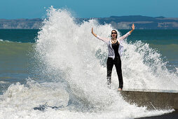 Fröhliche Frau steht am Meer in der Gischt einer Welle (MR), Napier, Hawke's Bay, Nordinsel, Neuseeland