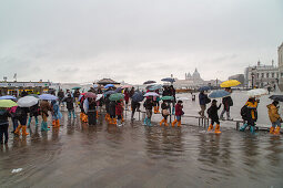Tourists with umbrellas and plastic covered shoes, rubber boots, wade through Acqua alta St Mark's Square, San Marco, high water caused by Sirocco and full moon, rain in Venice, Italy