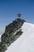 Two female alpinists holding on to the summit cross of Allalinhorn and waving, Pennine Alps, canton of Valais, Switzerland