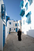 Woman walks along path of buildings with blue windows and white walls in artisan village, Sidi Bou Said, Tunis, Tunisia