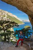 A young woman with trekking gear hikes through the rock arch Arcu su Feilau at the mountainous coast above the sea, Golfo di Orosei, Selvaggio Blu, Sardinia, Italy, Europe