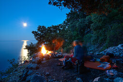 Junge Frau und junger Mann sitzen auf Isomatte am Lagerfeuer mit Vollmond auf einem Köhlerkreis über dem Meer, bei der Bucht Cala Biriola, Golfo di Orosei, Selvaggio Blu, Sardinien, Italien, Europa