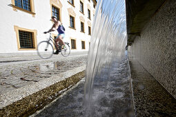 Junge Fahrradfahrerin fährt an einem Brunnen vorbei, Kempten, Bayern, Deutschland