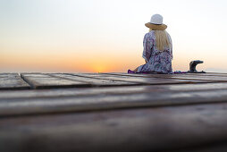 Young blond-haired woman sitting on the end of a long pier in the morning mood, enjoying the view of the sunrise. Playa de Muro beach, Alcudia, Mallorca, Balearic Islands, Spain