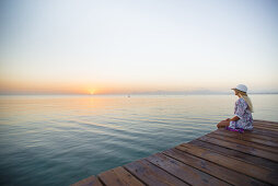 Young blond-haired woman sitting on the end of a long pier in the morning mood, enjoying the view of the sunrise. Playa de Muro beach, Alcudia, Mallorca, Balearic Islands, Spain