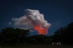 Blick auf eine sehr große Aschesäule des aktiven Vulkans Yasur bei Vollmond. Aschwolke wird unten von Lava ausgeleuchtet, oben vom Mond angestrahlt. Sternenhimmel, Wald und ein Haus mit ausgeleuchteten Fenstern in der Bildmitte, Nebel auf der Wiese im Vor