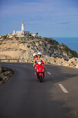Young couple riding a red Vespa scooter on a coastal road along the Cap de Formentor peninsula with Faro de Formentor lighthouse behind, Palma, Mallorca, Balearic Islands, Spain